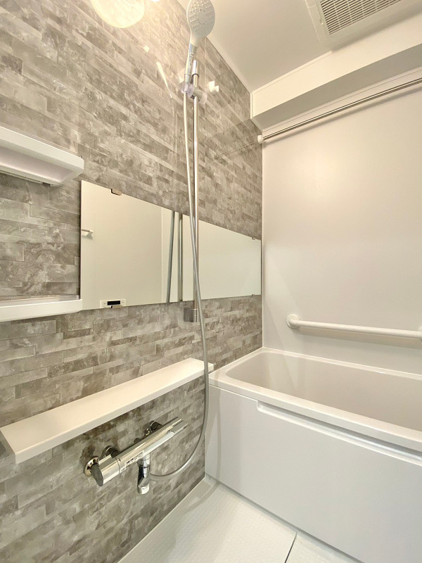 ヴェラハイツ新宿　バスルーム バスルームはグレーのレンガ調デザインパネルが施されたお洒落な空間✨モノトーン調で清潔感がありますね。バスタブの近くには手すりが設置され、入浴時の立ち座りを補助してくれますよ。浴室乾燥機や追い焚き機能などの便利な機能も搭載されています◎