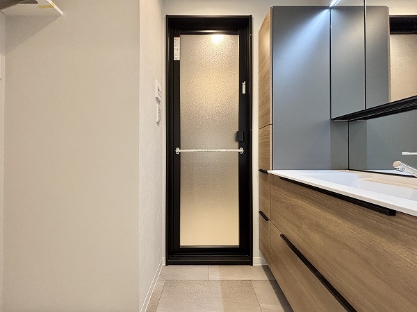 月島ホームズ_清潔感のあるパウダールームの写真 柔らかな色合いでまとめられたパウダールームは、落ち着きと清潔感にあふれる空間。洗面化粧台は3面鏡で、下部にも鏡があるので小さなお子さまでも身支度しやすく、家族みんなが使いやすい設計です。洗濯機上部の棚には洗剤やランドリーグッズをまとめて置けるため、家事動線もスムーズ。