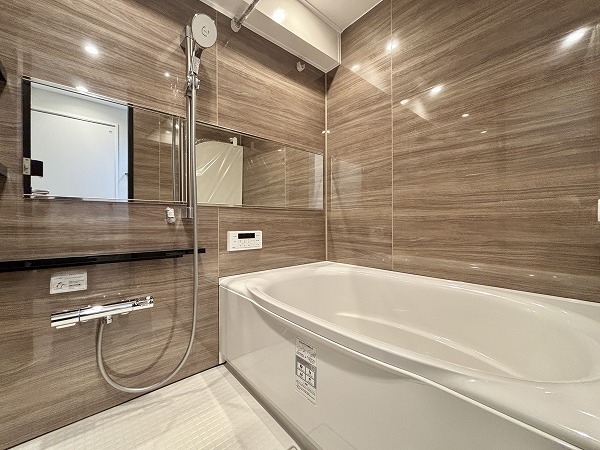 月島ホームズ_ブラウンを基調としたバスルームの写真 深みのあるブラウンパネルが落ち着いた高級感を演出するバスルーム。床には水はけの良い素材を採用しているため、入浴後もサッと乾き、カビの発生を防ぎます。丸みのあるバスタブにはオートバスと追い焚き機能を搭載しており、ご家族全員が温かいお風呂にゆったり浸かれますよ。